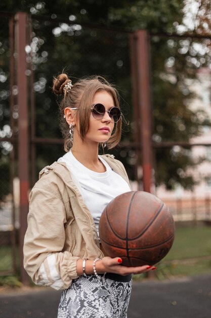 Elegante donna retrò con occhiali da sole vintage e acconciatura in abiti alla moda con giacca a vento Maglietta e leggings gioca con la palla sul campo da basket