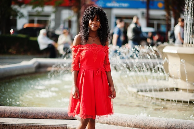 Elegante donna riccia afro francese alla moda posta al giorno d'autunno in abito rosso Modello femminile africano nero contro la fontana