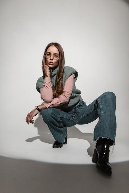 Elegante e affascinante bella giovane donna con occhiali vintage in abiti alla moda con una camicia rosa maglione lavorato a maglia e jeans larghi svasati seduto e in posa in studio Bella ragazza