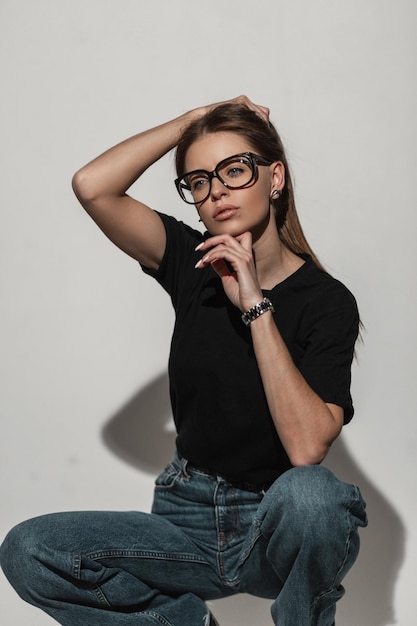 Elegante e bella giovane donna modello con occhiali vintage in maglietta nera alla moda e blue jeans accovacciati in studio Ragazza glamour