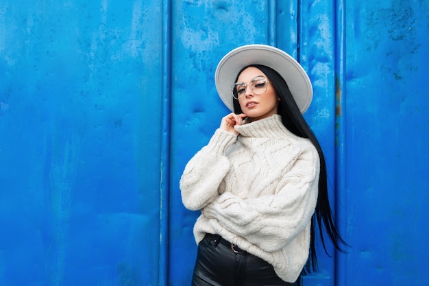 Elegante e bella ragazza con occhiali vintage e un cappello in un maglione lavorato a maglia alla moda si trova vicino a un muro di metallo blu sulla strada