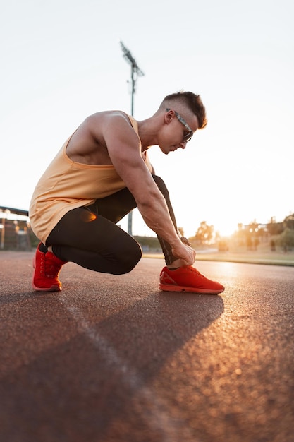 Elegante e bello sportivo giovane atleta con occhiali da sole in abbigliamento sportivo alla moda si siede e allaccia i lacci delle scarpe in scarpe da ginnastica rosse sullo stadio al tramonto Stile di vita sano e concetto di corsa