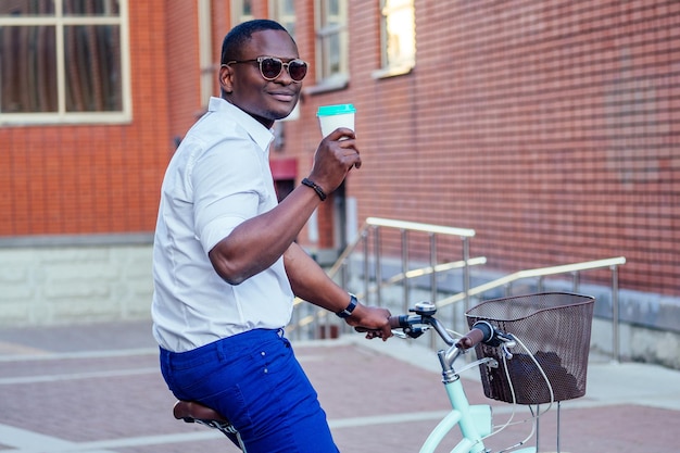 Elegante e giovane uomo afroamericano che tiene un bicchiere con caffè seduto su una bicicletta nel parco estivo