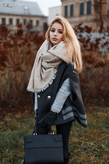 Elegante e moderna giovane donna in un maglione lavorato a maglia grigio in un cappotto nero con una sciarpa in jeans neri con una borsa di moda in pelle si trova vicino all'erba secca della città. Ragazza attraente.