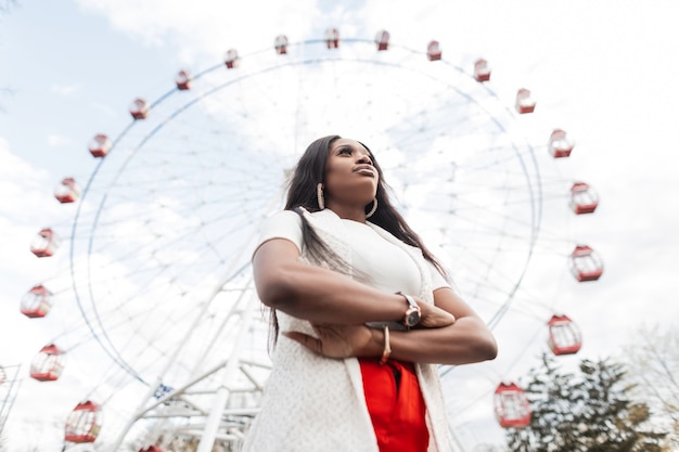 Elegante e moderna giovane donna nera sexy in abiti eleganti si erge contro il cielo e la ruota panoramica. La bella ragazza africana alla moda viaggia sulla città vicino al parco di divertimenti. Abbigliamento elegante per le donne.