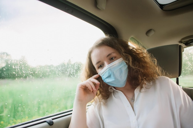 Elegante giovane donna carly in maschera medica seduta sul sedile posteriore dell'auto con sfondo con tramonto