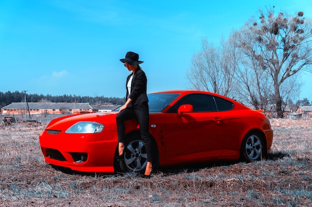 Elegante giovane donna con cappello a tesa larga siede su un'auto sportiva rossa nel campo