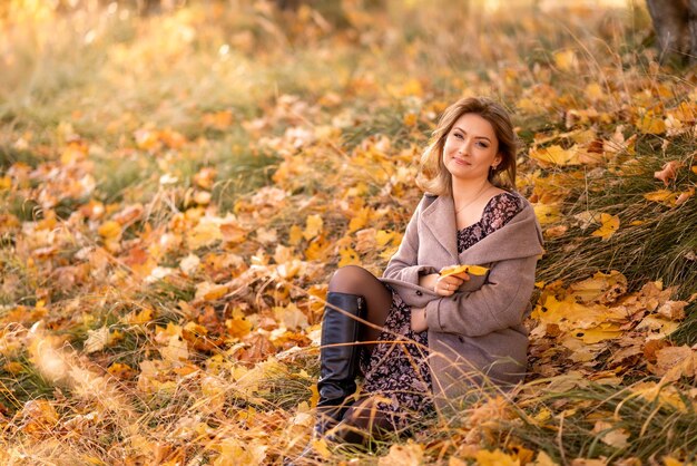 Elegante giovane donna con un cappotto grigio si siede su foglie gialle in un parco autunnale