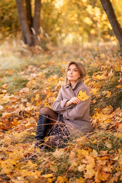 Elegante giovane donna con un cappotto grigio si siede su foglie gialle in un parco autunnale