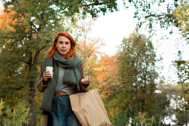 Elegante giovane donna dai capelli rossi in cappotto con caffè e borse della spesa artigianali che camminano nel parco cittadino in autunno