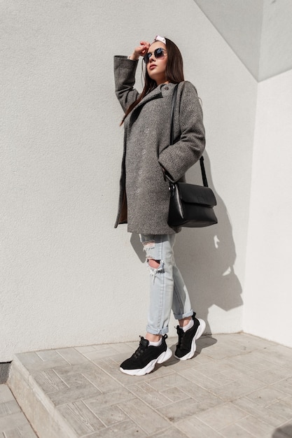 Elegante giovane donna in cappotto giovanile casual alla moda con borsa in scarpe da ginnastica con bandana in occhiali da sole sta riposando in una giornata di sole vicino al muro vintage sulla strada. Attraente ragazza urbana e sole splendente.