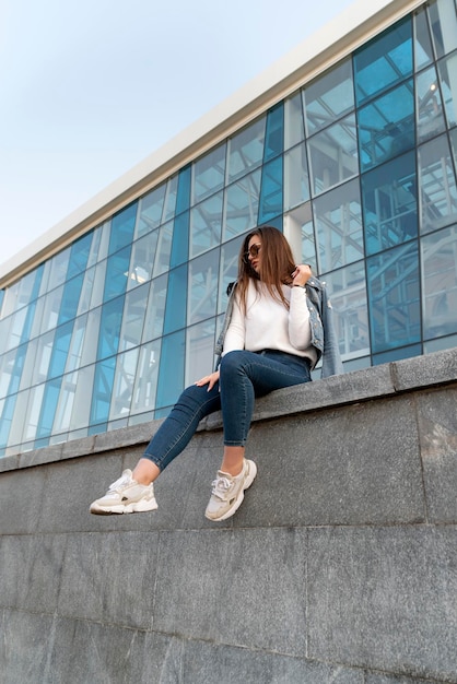 Elegante giovane donna in occhiali da sole seduta sulla strada della città Elegante donna che riposa accanto al moderno edificio Verticale