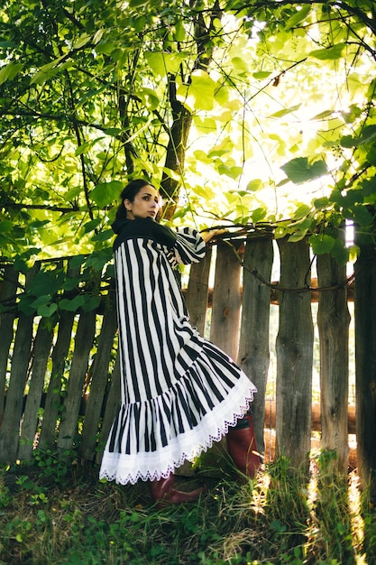 Elegante giovane donna indiana in abito bianco e nero su uno sfondo di alberi verdi e recinto di legno invecchiato Boho donna rilassante in campagna semplice stile di vita lento Immagine atmosferica