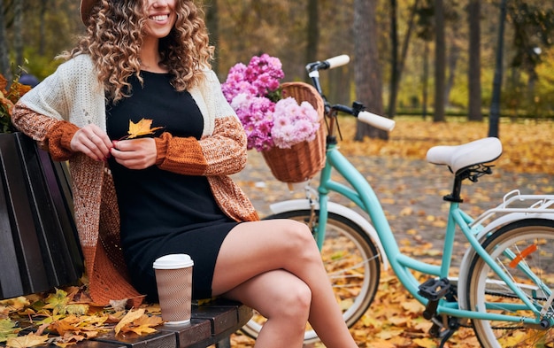 Elegante giovane donna seduta su una panchina vicino alla bicicletta nel parco autunnale