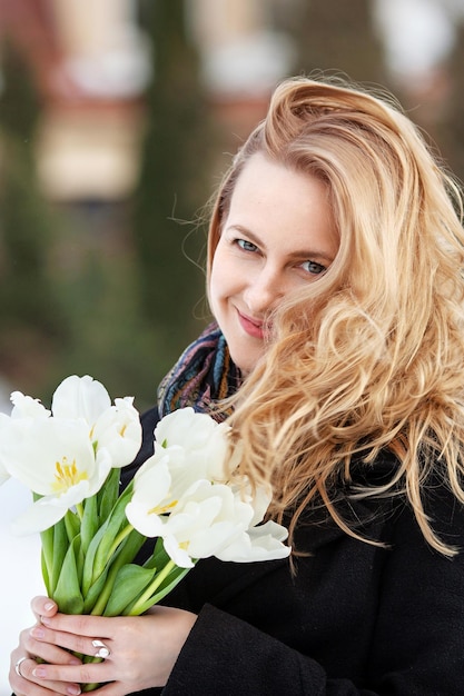 Elegante giovane donna sorridente con lunghi capelli biondi in un cappotto nero posa in città