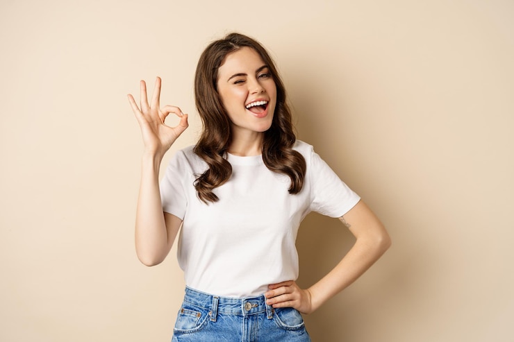 Foto elegante giovane donna sorridente soddisfatta, mostrando ok, ok firmare in approvazione, consigliando, in piedi su sfondo beige.