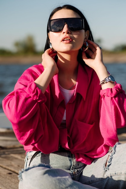 Elegante giovane donna vestita in casual in occhiali da sole seduta sul molo di legno in una giornata di sole