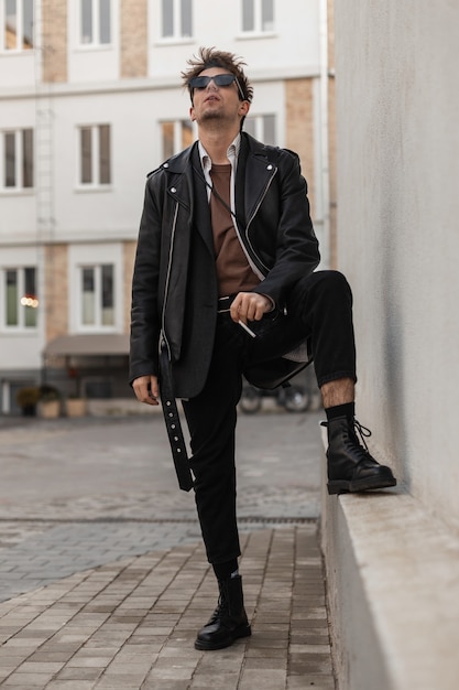 Elegante giovane hipster in occhiali da sole scuri alla moda in una giacca di pelle nera in jeans in stivali cool si alza e fuma vicino a un muro vintage. Bel modello di moda ragazzo americano su una pausa fumo all'aperto