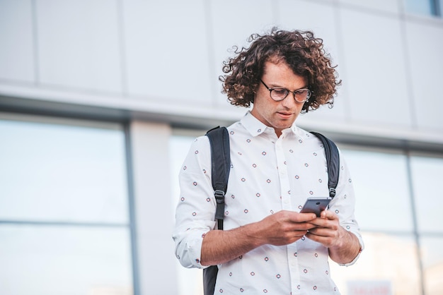 Elegante giovane maschio caucasico serio che indossa occhiali alla moda e camicia guardando lo schermo del suo smartphone in procinto di rispondere ad alcune e-mail Persone stile di vita e comunicazione tecnologica moderna