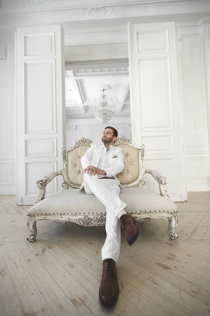 Elegante giovane uomo bello con la barba in un abito classico bianco. Gli interni dell'hotel. Studio fotografico. Divano. Finestra. Sedia. Lampadario