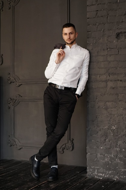 Elegante giovane uomo d'affari in camicia bianca è in possesso di una giacca sul dito in piedi contro il muro di mattoni Foto verticale