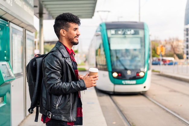 Elegante giovane uomo latino con un caffè da asporto in mano aspetta alla fermata del trasporto pubblico l'arrivo del tram, concetto di stile di vita urbano