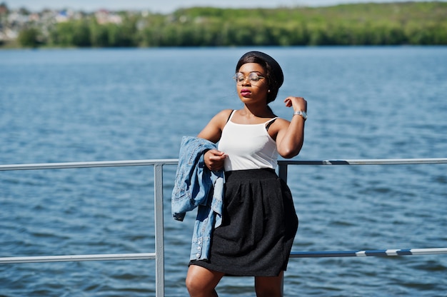 Elegante modello afro-americano in cappello occhiali, giacca di jeans e gonna nera poste all'aperto sul molo contro il lago.