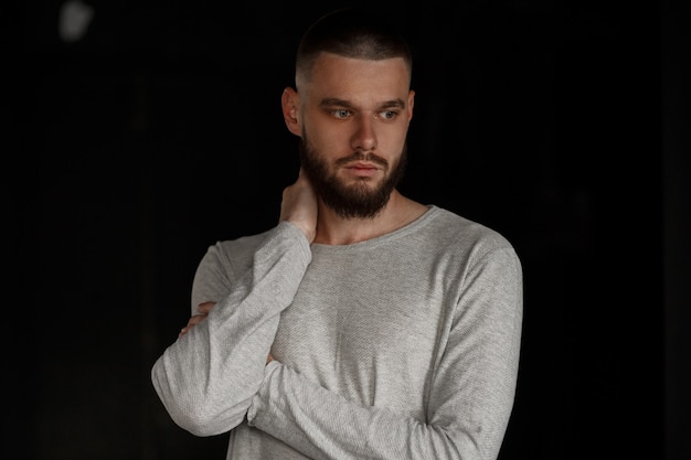 Elegante modello bello uomo con la barba in maglietta grigia sul muro nero