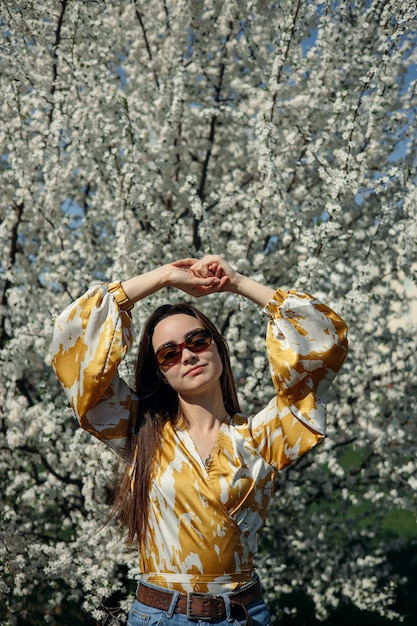 Elegante modello femminile in camicetta e occhiali da sole in piedi contro alberi con fiori nel giardino fiorito