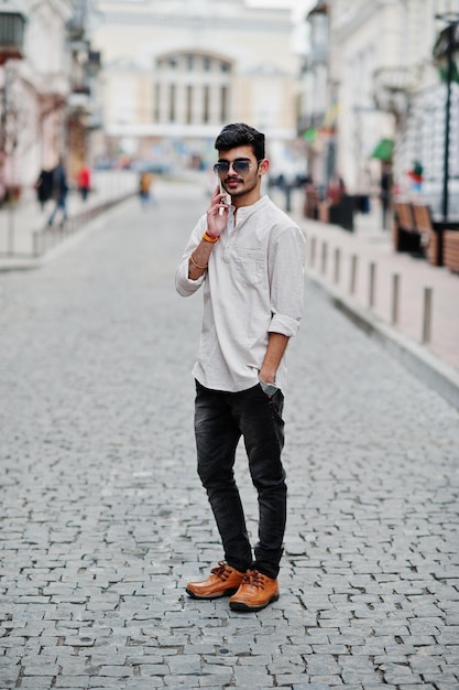 Elegante modello indiano uomo in abiti casual e occhiali da sole posato all'aperto in strada dell'India e parlando al telefono cellulare