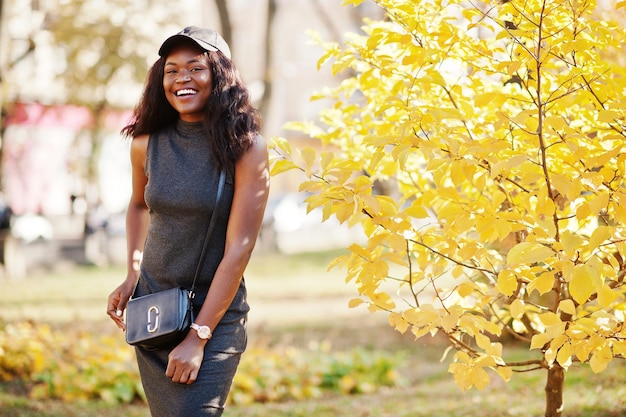 Elegante ragazza afroamericana in tunica grigia borsa a tracolla e cappuccio poste in una soleggiata giornata autunnale contro le foglie gialle Donna modello Africa