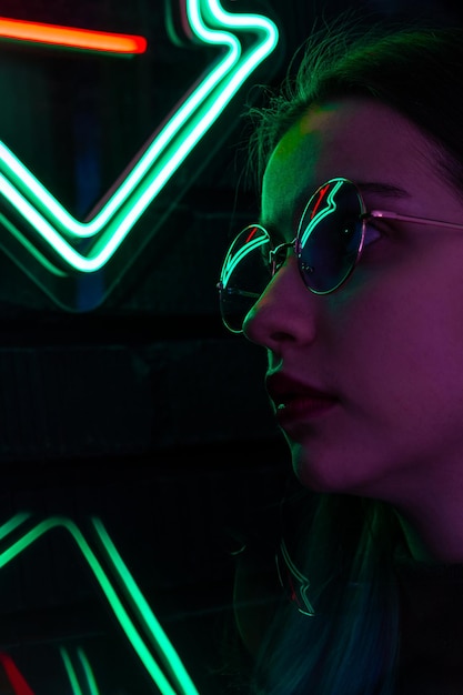 Elegante ragazza alla moda con gli occhiali con la riflessione guarda in alto sulla strada con l'illuminazione al neon del bar della città e del concetto musicale
