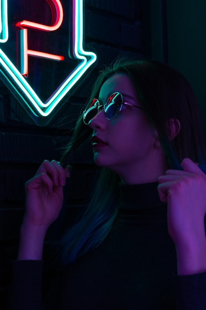 Elegante ragazza alla moda con gli occhiali con la riflessione guarda in alto sulla strada con l'illuminazione al neon del bar della città e del concetto musicale