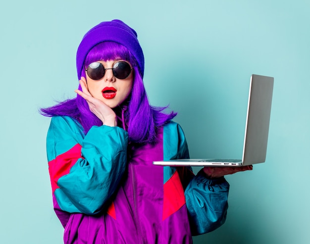 Elegante ragazza bianca con capelli viola e tuta con il computer portatile sulla parete blu