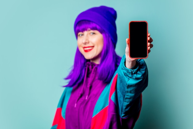 Elegante ragazza bianca con i capelli viola e tuta utilizzando il telefono cellulare sulla parete blu