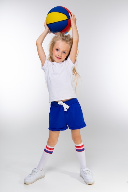 Elegante ragazza bionda in pantaloncini sportivi, una t-shirt e scarpe da ginnastica con un pallone da basket su un muro bianco