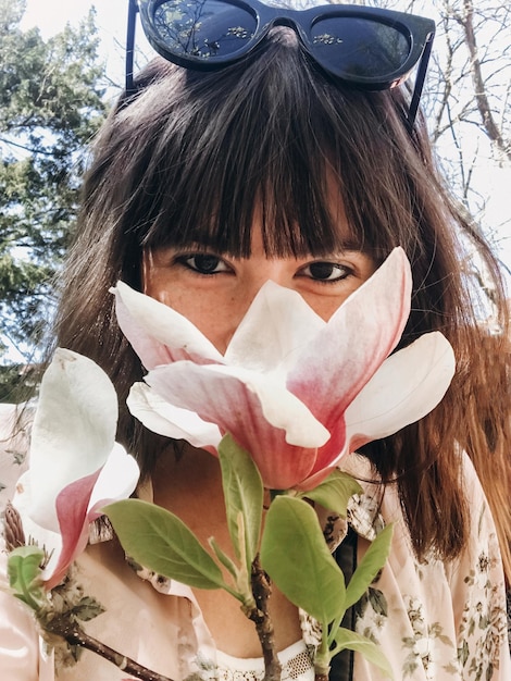 Elegante ragazza boho che fa selfie con bellissimi fiori di magnolia rosa sul ramo Ragazza hipster che odora e posa con fiori di magnolia sull'albero nel soleggiato giardino botanico Foto del telefono Ciao primavera