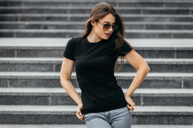 Elegante ragazza bruna che indossa maglietta nera e occhiali in posa contro lo stile di abbigliamento urbano di strada
