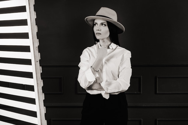 Elegante ragazza bruna con una camicia bianca e un cappello beige su uno sfondo scuro. foto in bianco e nero