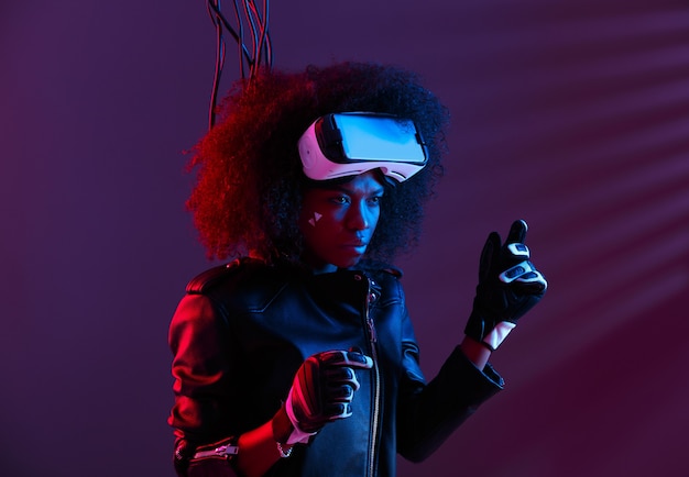 Elegante ragazza dai capelli scuri riccia vestita con giacca di pelle nera e guanti indossa gli occhiali per realtà virtuale sulla testa nello studio buio con luce al neon. Personaggio cyberpunk