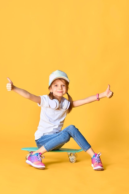 Elegante ragazza divertente che indossa t-shirt bianca, jeans blu e scarpe da ginnastica, seduto su skateboard su uno spazio giallo e alza le mani e il pollice in alto emotivamente