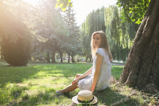 Elegante ragazza hipster con un cappello di paglia si sta godendo il sole all'aperto Donna felice seduta in abito blu vicino all'albero Concetto di vacanza di viaggio Stile di vita estivo Sfumature sugli alberi Bei capelli