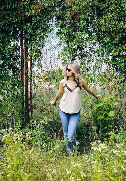 Elegante ragazza hipster è in piedi vicino al muro di foglie di edera all'aperto Servizio fotografico alla moda per le giovani donne Capelli corti biondi e occhiali fantastici