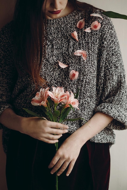 Elegante ragazza hipster in cappello con fiori rosa e petali su maglione che cresce boho donna che tiene bella alstroemeria nelle mani in primavera mattina ritratto femminile sensuale creativo