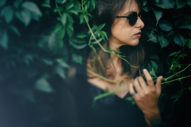 Elegante ragazza hipster in posa al momento atmosferico delle foglie verdi Ritratto di donna cool alla moda in occhiali da sole neri che si rilassano nel parco Immagine creativa di messa a fuoco selettiva Spazio per il testo