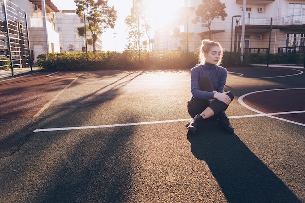 Elegante ragazza modello bionda in maglione blu si siede per terra in un'area sportiva all'aperto al sole