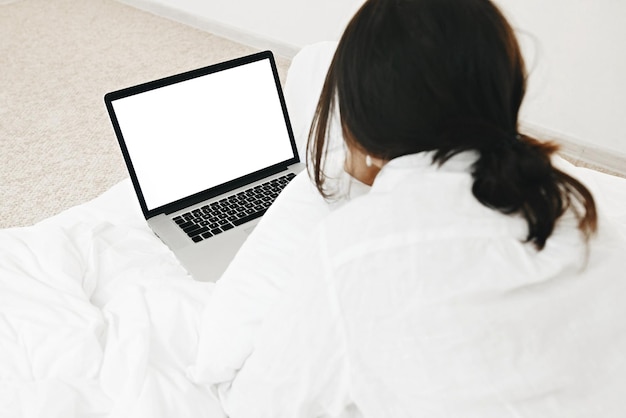 Elegante ragazza sdraiata sul letto con un computer portatile con schermo vuoto in camera bianca moderna Giovane donna che guarda un display vuoto che fa shopping online o lavora online da casa modello Freelance