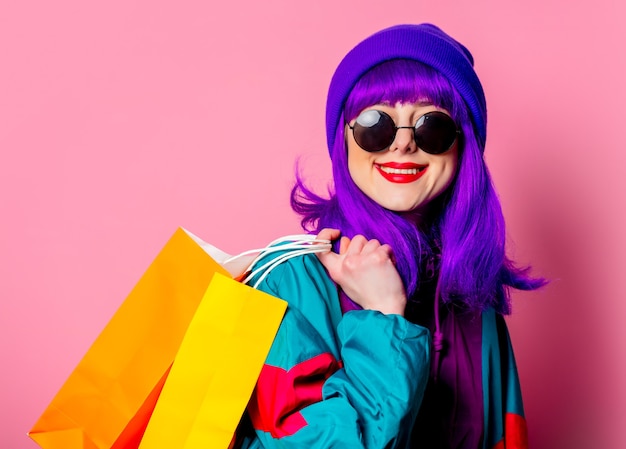 Elegante ragazza shopaholic in tuta da ginnastica e occhiali da sole tenere borse della spesa sulla parete rosa