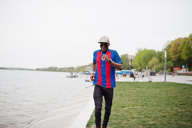 Elegante ragazzo afroamericano che corre contro il lago indossa una maglietta da calcio e occhiali da sole Ritratto di uomo sportivo nero