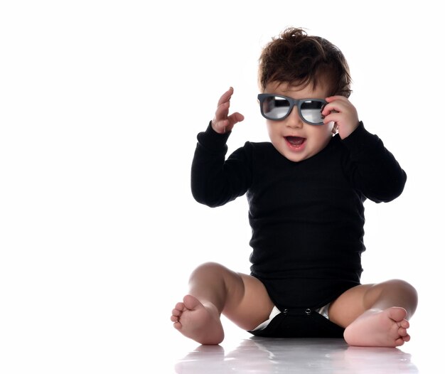 Elegante ragazzo alla moda in tuta nera occhiali da sole seduto sul pavimento Sorridente positivo bambino bambino Ritratto isolato girato su uno sfondo bianco per studio Stile e moda infanzia felici